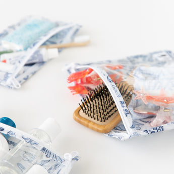 Close-up of Roamio by Bumkins Clear Travel Bag 3 Pack: Shells on a white background with a hairbrush, hair accessories, & toiletries.