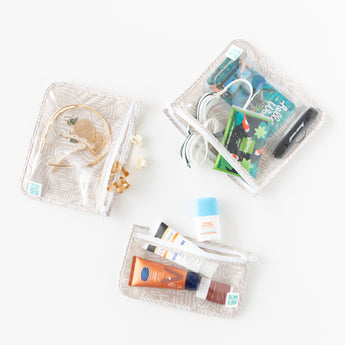 Bumkins Clear Travel Bag 3-Pack: Wander  is neatly arranged on a white background holding toiletries, hair accessories, books, & goggles.