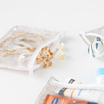 Close-up of Bumkins Clear Travel Bag 3-Pack: Wander on a white background with toiletries & hair accessories peeking out.