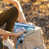 A person places bags from Roamio by Bumkins The Commuter Deluxe 5-Piece Set: Denim On Denim in a backpack on rocky ground.