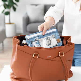 A person places a Roamio by Bumkins Clear Travel Bag 3 Pack: Crushed Denim with a book and cable in a brown purse.