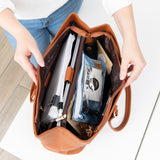 A person opens a brown purse on a white table, revealing notebooks and a Roamio by Bumkins Clear Travel Bag 3 Pack: Crushed Denim inside.