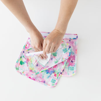 Hands holding a floral bag from the Bumkins Clear Travel Bag 3-Pack: Watercolor, another similar bag underneath on a white background.