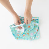 Hands place a small item in a TSA-approved Bumkins Clear Travel Bag from the Ocean Life 3-Pack on a white surface.