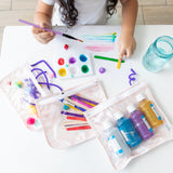 A girl paints at a white table near bags from Bumkins The Chaos Coordinator Essential 4-Piece Set: Lace filled with art supplies.