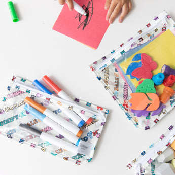 A child draws near bags from Bumkins The Chaos Coordinator Deluxe 5-Piece Set: Disney Mickey & Friends Varsity with art supplies on a table.