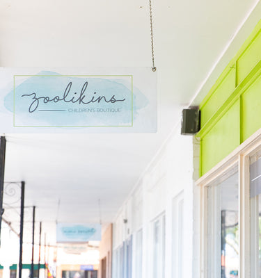 Sign hanging under a white porch ceiling reads Zoolikins Childrens Boutique. The building features a bright lime-green accent wall and blue shutters. Ornate metal brackets support the porch roof, and a tree-lined street is visible on the side.