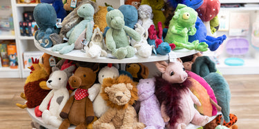 A display shelf filled with colorful stuffed animals, including teddy bears, dinosaurs, and a pig, in a toy store setting. The toys are arranged on two white circular tiers.