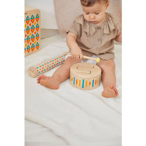 A baby on a soft blanket explores PlanToys Solid Drum II and a mallet, boosting motor skills. A tall patterned toy is nearby.