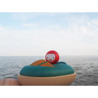 A PlanToys Wooden Speed Boat Bath Toy features a red-headed, smiling figurine in a sustainable boat against the sea and clouds.