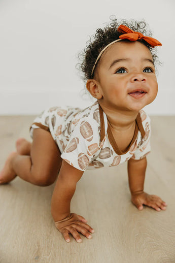 A baby smiles upward while crawling on a wooden floor in a Copper Pearl Blitz bodysuit and an orange bow headband.
