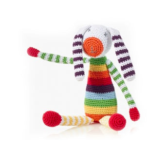 The Hand Knit Rainbow Bunny by Pebble is crocheted from natural materials, with multicolored stripes, long ears, limbs, and a raised arm.