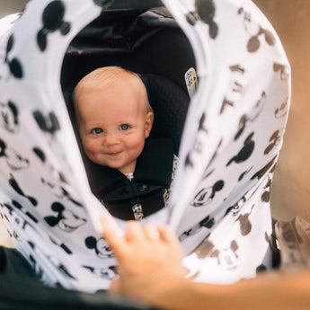 A smiling baby in a stroller with a Milk Snob Cover Disneys Mickey Sketch, gently pulled back by an adult hand.