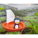 A toy penguin from PlanToys sails in a PlanWood orange boat with a white sail on a pond surrounded by lush leaves.