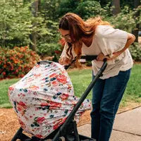 A woman with long hair leans over a stroller, using a Milk Snob Cover French Floral, in a serene park setting.
