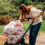 A woman with long hair leans over a stroller, using a Milk Snob Cover French Floral, in a serene park setting.