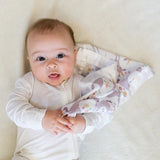 Baby in a white onesie cuddling Lulujo Inc.s colorful, breathable muslin Unicorn Security Blanket on a light blanket.