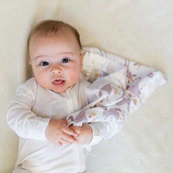 Baby in a white onesie cuddling Lulujo Inc.s colorful, breathable muslin Unicorn Security Blanket on a light blanket.