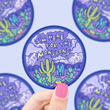 A hand holds Turtle Soup’s Here For the Monsoon vinyl sticker with a desert scene of cacti and lightning.