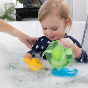 A toddler giggles, splashing in a bubble bath with Dimpl Splash by Fat Brain Toy Co, enhancing fine motor skills with a caregiver.