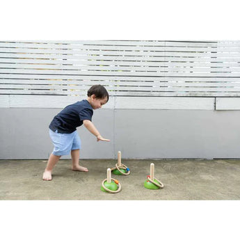 A child practices motor skills playing PlanToys Meadow Ring Toss on concrete, aiming at pegs near a white fence.