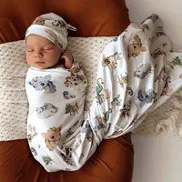 A baby sleeps on a brown cushion, wrapped in the Snuggle Hunny Organic Jersey Wrap & Beanie Set, Dragon.