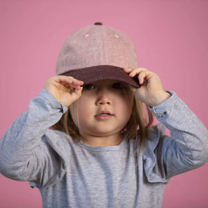 A child in a gray shirt adjusts their Lil Hat, Jam from Storied Hats against a pink background.