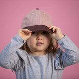 A child in a gray shirt adjusts their Lil Hat, Jam from Storied Hats against a pink background.