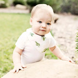 A baby in a white outfit and Bebe Au Laits Saguaro Bandana Bib stands by a rock, with grass and blurred greenery in the background.