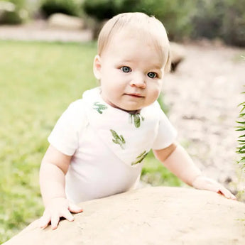 A baby in a white outfit and Bebe Au Laits Saguaro Bandana Bib stands by a rock, with grass and blurred greenery in the background.