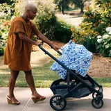 Person in an orange dress pushes stroller with a Milk Snob Cover Disneys Stitch, walking through a garden.