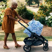 Person in an orange dress pushes stroller with a Milk Snob Cover Disneys Stitch, walking through a garden.