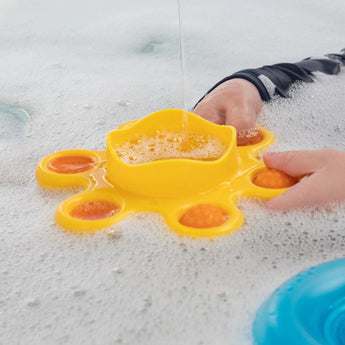 Hands engage with the Dimpl Splash by Fat Brain Toy Co in soapy water; its yellow design enhances fine motor skills through sensory play.