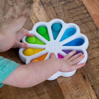 A child plays with the Fat Brain Toy Cos Dimpl Digits, a colorful flower-shaped sensory toy, on a wooden floor.