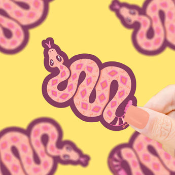 A hand with painted nails holds Turtle Soups Vinyl Sticker, Slithering Rattlesnake on a yellow background.