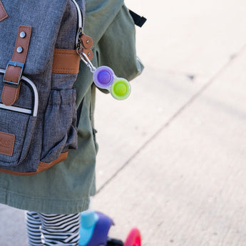 A child in a green jacket holds a backpack with a Fat Brain Toy Cos Simpl Dimpl. A colorful scooter is nearby on the sidewalk.