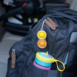 Gray backpack on car seat with colorful sippy cup in side pocket and Fat Brain Toy Cos clear Simpl Dimpl attached to the zipper.