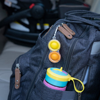 Gray backpack on car seat with colorful sippy cup in side pocket and Fat Brain Toy Cos clear Simpl Dimpl attached to the zipper.