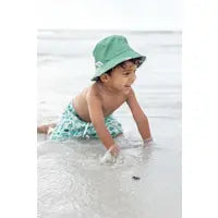 A baby in an Emerson and Friends Sea Turtles Reversible Bucket Hat plays happily in the sand by the beach.