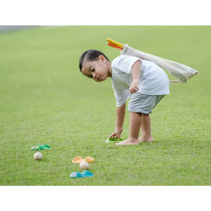 A child plays on the grass with a PlanToys Mini Golf Set, arranging colorful cones and balls, toy bag slung over their shoulder.