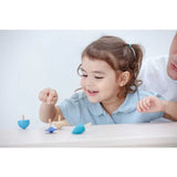 A young child smiles while playing with PlanToys Spinning Tops, enhancing fine motor skills, as an adult supervises.