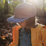 A child in a yellow raincoat wears Storied Hats Lil Hat, Khaki Southwest, gazing down in a sunlit forest.