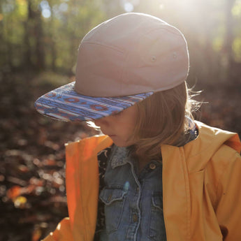 A child in a yellow raincoat wears Storied Hats Lil Hat, Khaki Southwest, gazing down in a sunlit forest.