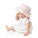 Baby wearing Bumkins Sea Unicorn pink sun hat (UPF 50+), white dress, sits sideways with fingers in mouth.