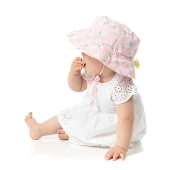 Baby wearing Bumkins Sea Unicorn pink sun hat (UPF 50+), white dress, sits sideways with fingers in mouth.