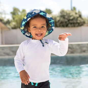 A gleeful toddler in swimwear and a Bumkins Deep Sea sun hat (UPF 50+) waves happily by the pool.