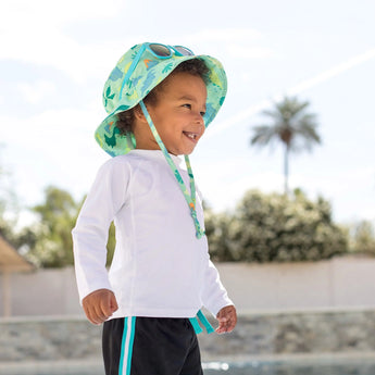 A toddler in a Bumkins Sun Hat with Green Dinosaurs smiles outdoors by water, framed against palm trees and a clear sky.