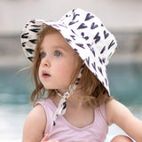 A young child wearing the Bumkins Sun Hat: Hearts with UPF 50+ sits poolside, gazing upward. The hat is white with black hearts.