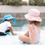 Two kids in UPF 50+ Bumkins sea unicorn sun hats and swimsuits sit by the pool. One hat is blue; the other is pink with yellow accents.
