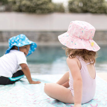 Two kids in UPF 50+ Bumkins sea unicorn sun hats and swimsuits sit by the pool. One hat is blue; the other is pink with yellow accents.